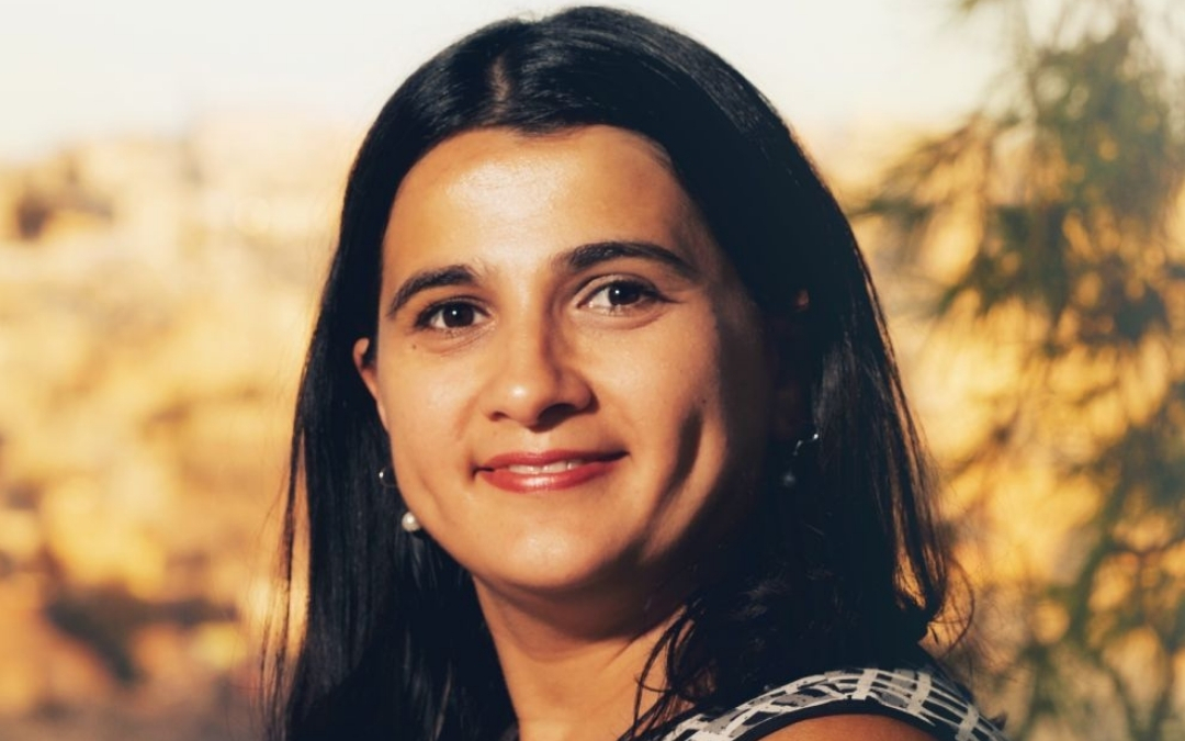 Author Natasha Tynes wears a patterned sleeveless top and pearl earrings, her long dark hair framing her face as she gazes softly toward the camera. The warm, blurred background suggests an outdoor setting at sunset, casting a gentle glow across her features and creating a calm, inviting portrait.