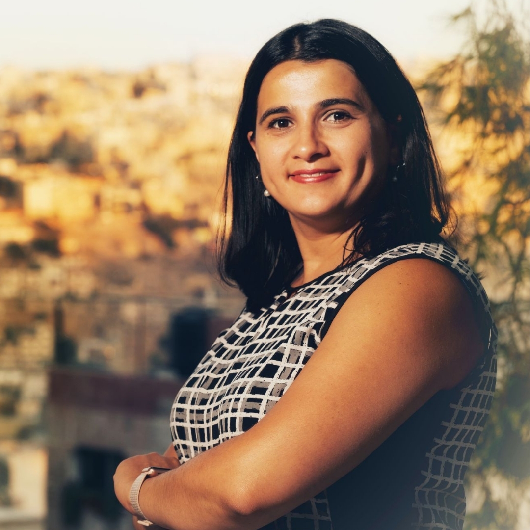 Author Natasha Tynes wears a patterned sleeveless top and pearl earrings, her long dark hair framing her face as she gazes softly toward the camera. The warm, blurred background suggests an outdoor setting at sunset, casting a gentle glow across her features and creating a calm, inviting portrait.