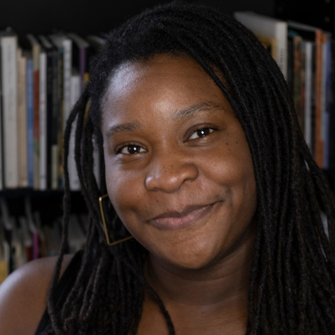 Maya Marshall 1x1 Author Maya Marshall stands in front of a bookshelf filled with a variety of books. Her long hair frames her face as she looks toward the camera, with the warm, book-lined setting suggesting a creative and literary environment.