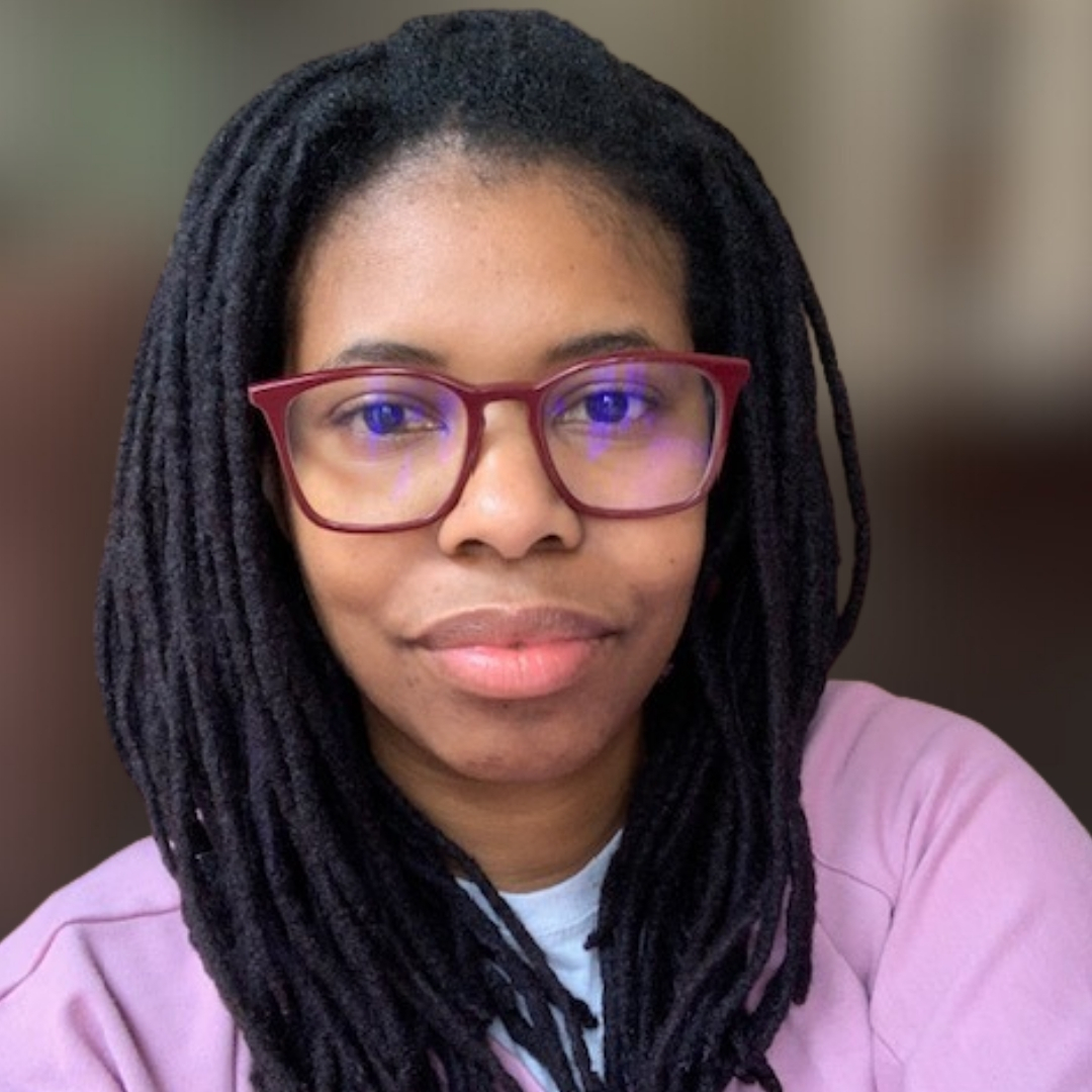 Alexis Laryea Color photo of Alexis Laryea indoors, wearing red-framed glasses and a light purple top. Her long, thick, twisted hair frames her face as she sits in a relaxed pose. The softly blurred background includes a partially open closet and shelves, suggesting a casual, home-like setting. Natural light highlights her features and adds warmth to the scene.