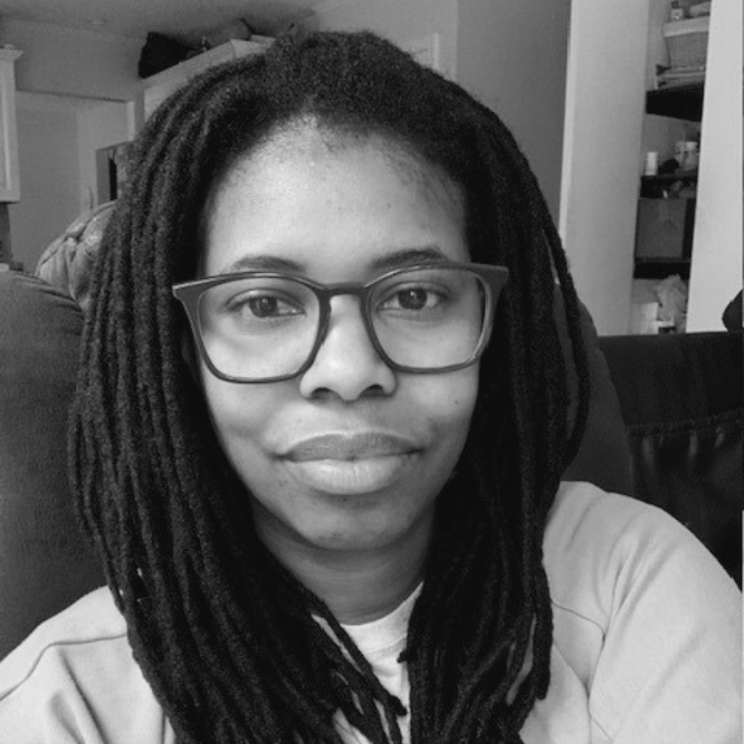 Black-and-white photo of Alexis Laryea indoors, wearing glasses and a light-colored top. She has long, thick, twisted hair and sits in a relaxed pose, with a partially open closet and shelves visible in the background.
