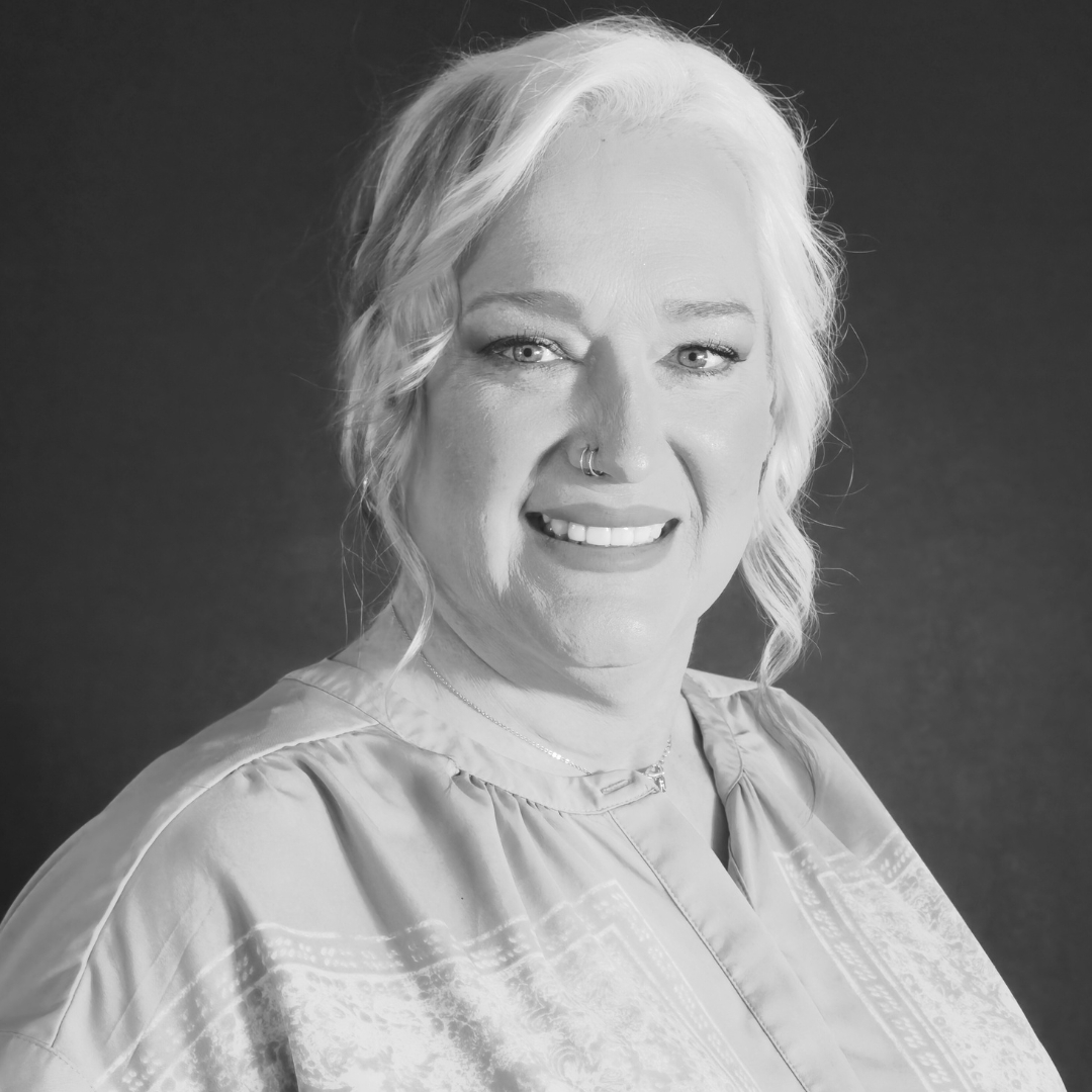 Black-and-white portrait of Alleria Stanley, shown from the shoulders up. She has light wavy hair that falls past her shoulders, a nose ring, and wears a patterned blouse. Her expression is calm and direct, and the dark background emphasizes the contours of her face and the texture of her hair.