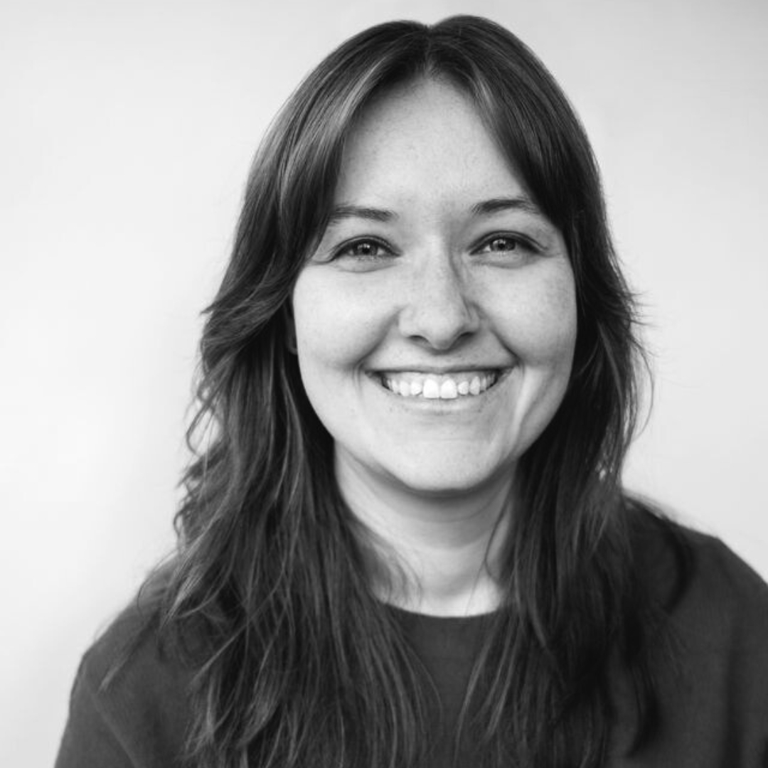 Black-and-white portrait of Amelia Bane, shown from the shoulders up, facing the camera with a gentle smile. She has long, dark hair and wears a dark top against a plain, light background. The image is evenly lit and emphasizes her facial expression and features.