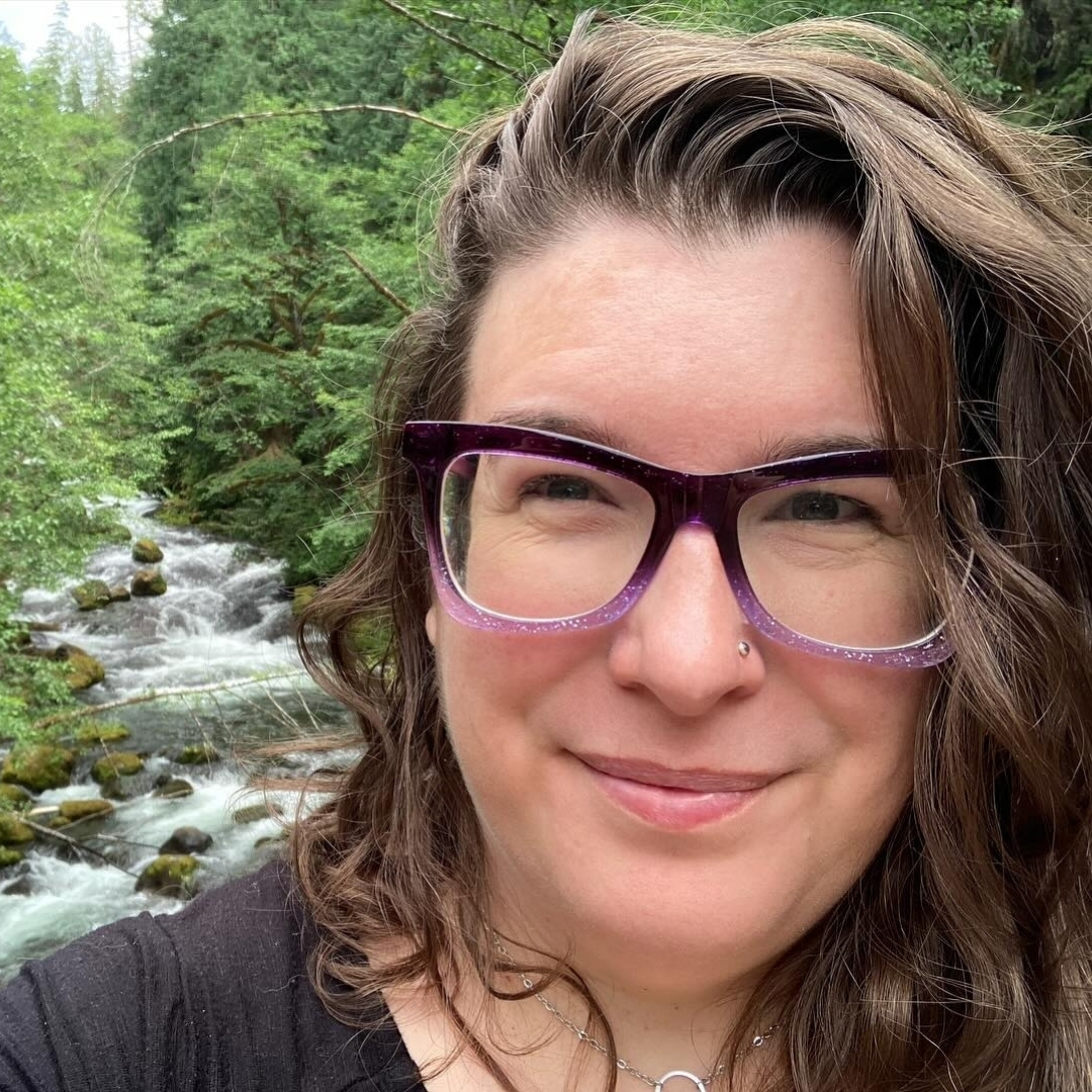 Angie Ebba Artist Angie Ebba, with long wavy hair, purple glasses, and a nose stud, smiles warmly at the camera. Behind her, a lush forest surrounds a rocky stream, with dense green trees and flowing water creating a peaceful, natural backdrop.