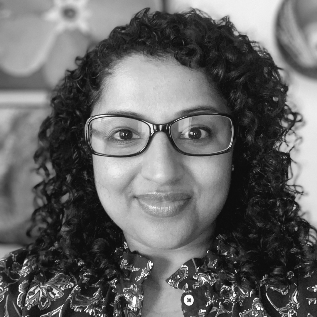 Black-and-white portrait of Ashy Palliparambil, a person with curly hair, glasses, and a patterned shirt, facing the camera with a gentle smile. The background is softly blurred, featuring abstract artwork and decorative elements including a flower and a circular object.