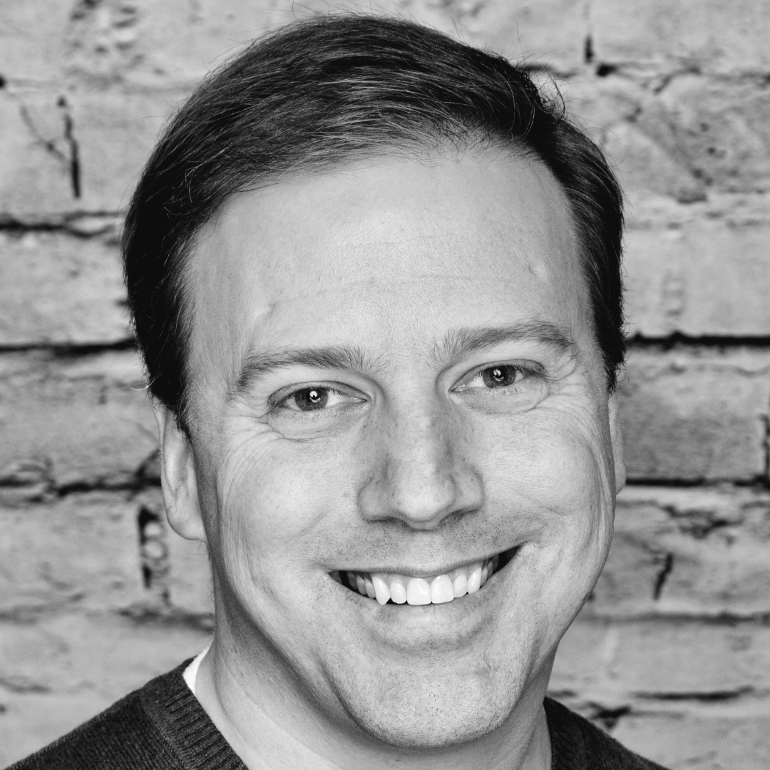 Black-and-white headshot of Ben Weakley smiling in front of a brick wall. The image is closely cropped to highlight his facial features and expression, with textured bricks providing contrast in the background.