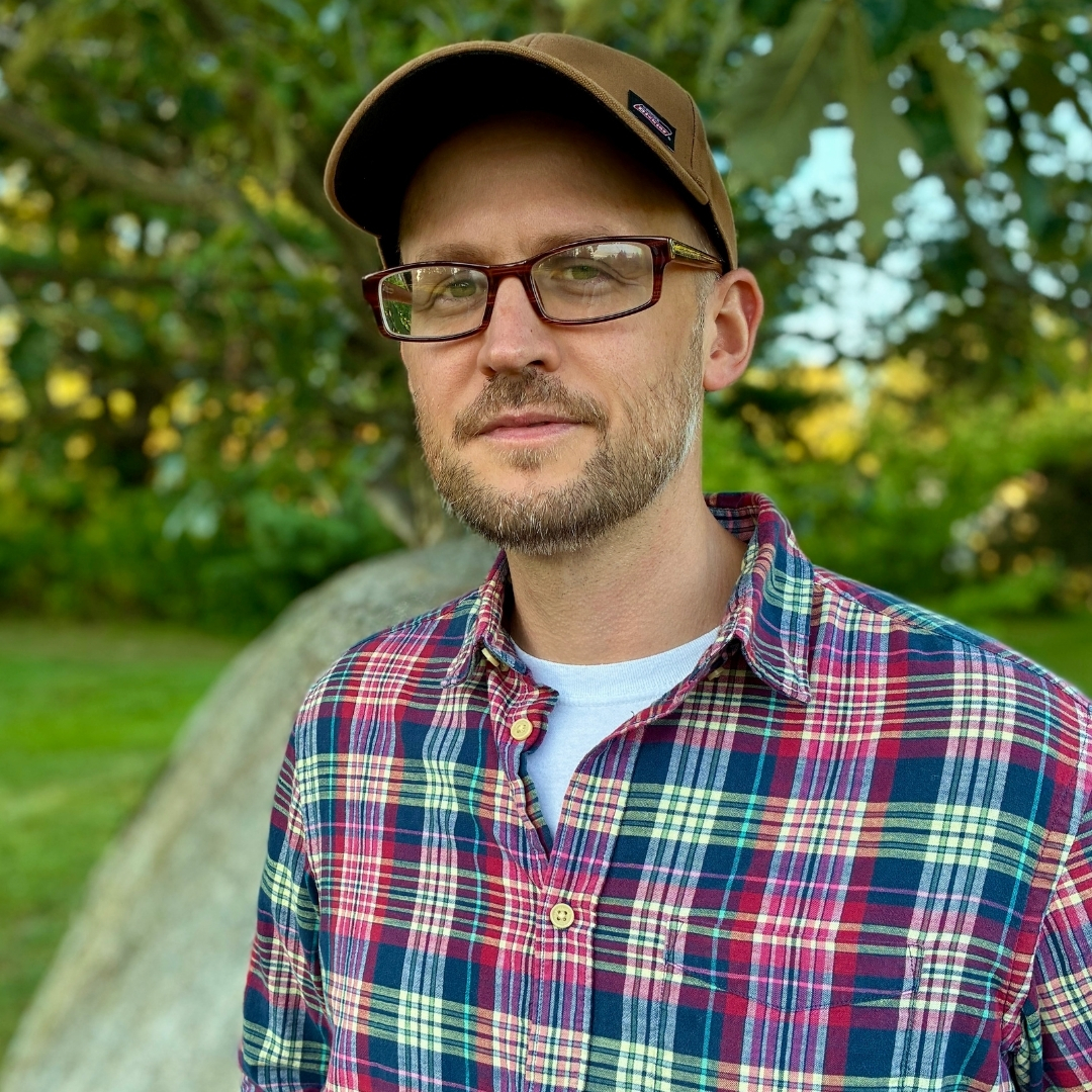 Kevin Basl Color portrait of Kevin Basl standing outdoors in front of a large tree with green foliage. He wears a brown cap, glasses, and a plaid shirt with red, blue, and yellow tones over a white undershirt. He looks slightly to the side with a calm expression. The background includes grass and trees, suggesting a peaceful park setting in natural light.