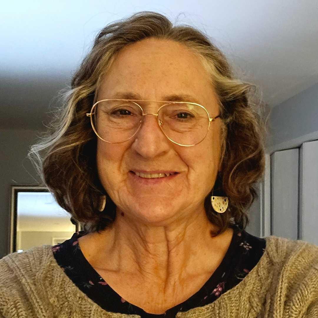 Liz Lockett - Headshot Portrait of Liz Lockett, shown from the shoulders up in a softly lit room with white walls and a mirror in the background. She has shoulder-length curly hair, wears glasses, and distinctive half-circle earrings with a speckled design. Her outfit includes a beige knitted sweater layered over a dark floral top, creating a warm and approachable look.