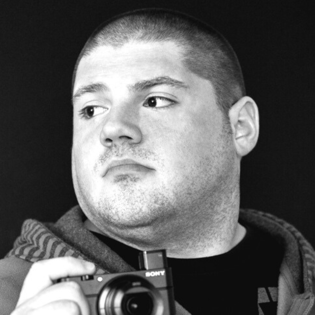 Black-and-white portrait of Rob Haney holding a Sony digital camera, looking slightly to the side. Rob has a closely shaved haircut and wears a hooded garment with a striped inner lining. The solid black background emphasizes Rob's facial features and the camera’s details.