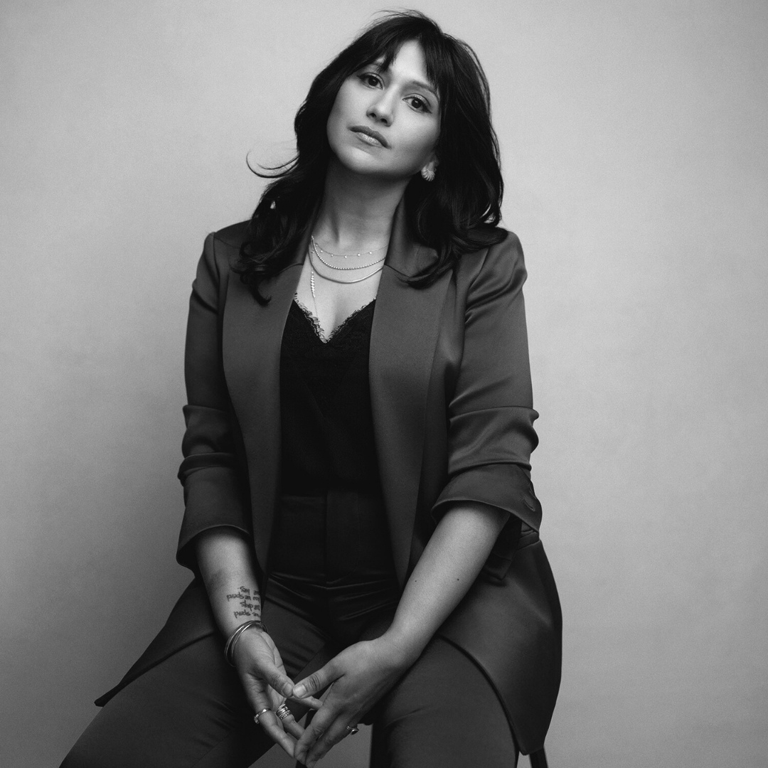 Black‑and‑white portrait of Seema Reza, Mission Belonging’s board chair, co‑founder, and CEO. She is seated against a plain backdrop, wearing a tailored suit over a lace‑trimmed top with layered jewelry. Her expression is calm and composed, and a forearm tattoo is visible, contributing to the image’s refined, monochromatic aesthetic.