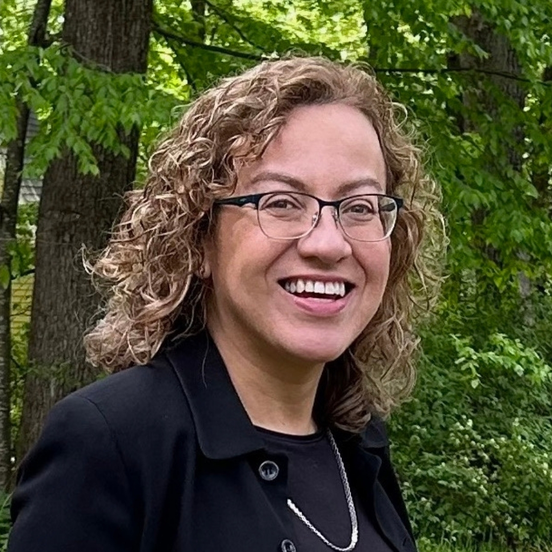 Luisa Caycedo-Kimura, wearing glasses and a dark jacket, smiles outdoors in a wooded area. Tall green trees fill the background, creating a lush forest setting. The lighting is soft and natural, capturing a candid, peaceful moment. (Photo by Aaron Caycedo-Kimura)
