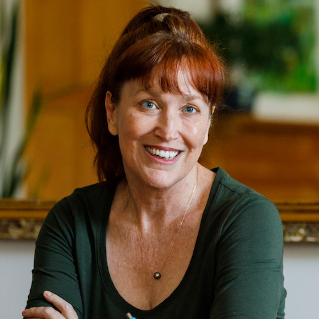 Allison K Williams Portrait-1080x1080 Portrait of author Allison K. Williams seated at a wooden table, smiling as she writes in an open notebook with a blue pen. She has red hair, wears a dark green top and a small pendant necklace, and is surrounded by a warm indoor setting with framed artwork and greenery in the background.