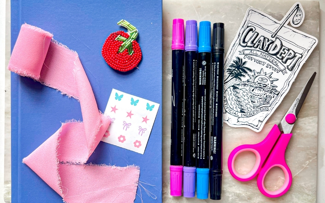A flat lay of crafting supplies arranged for Leslie Hidalgo’s Junk Journaling workshop. On the left, a closed blue book sits beneath a frayed pink ribbon, topped with a red beaded tomato-shaped pin and a small sheet of pastel stickers featuring butterflies, stars, bows, and flowers. On the right, four dual‑tip markers in pink, purple, blue, and black are lined up beside pink‑handled scissors and a black‑and‑white illustrated cutout labeled “CLAYDEPT All Natural Pottery Stuff.” The items rest on a soft, textured surface, creating a colorful, inviting setup for creative journaling.