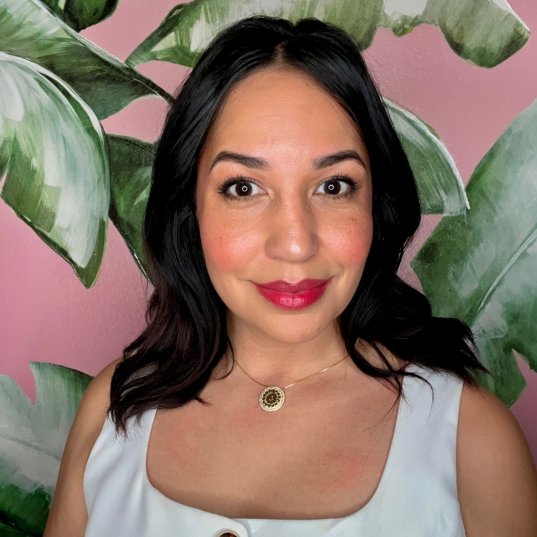 Leslie Hidalgo Portait Portrait of Leslie Hidalgo standing in front of a pink wall painted with large green and white leaves. She has dark hair, bright pink lipstick, and wears a white top with a circular pendant necklace. The vibrant background creates a bold contrast with her calm, confident expression.