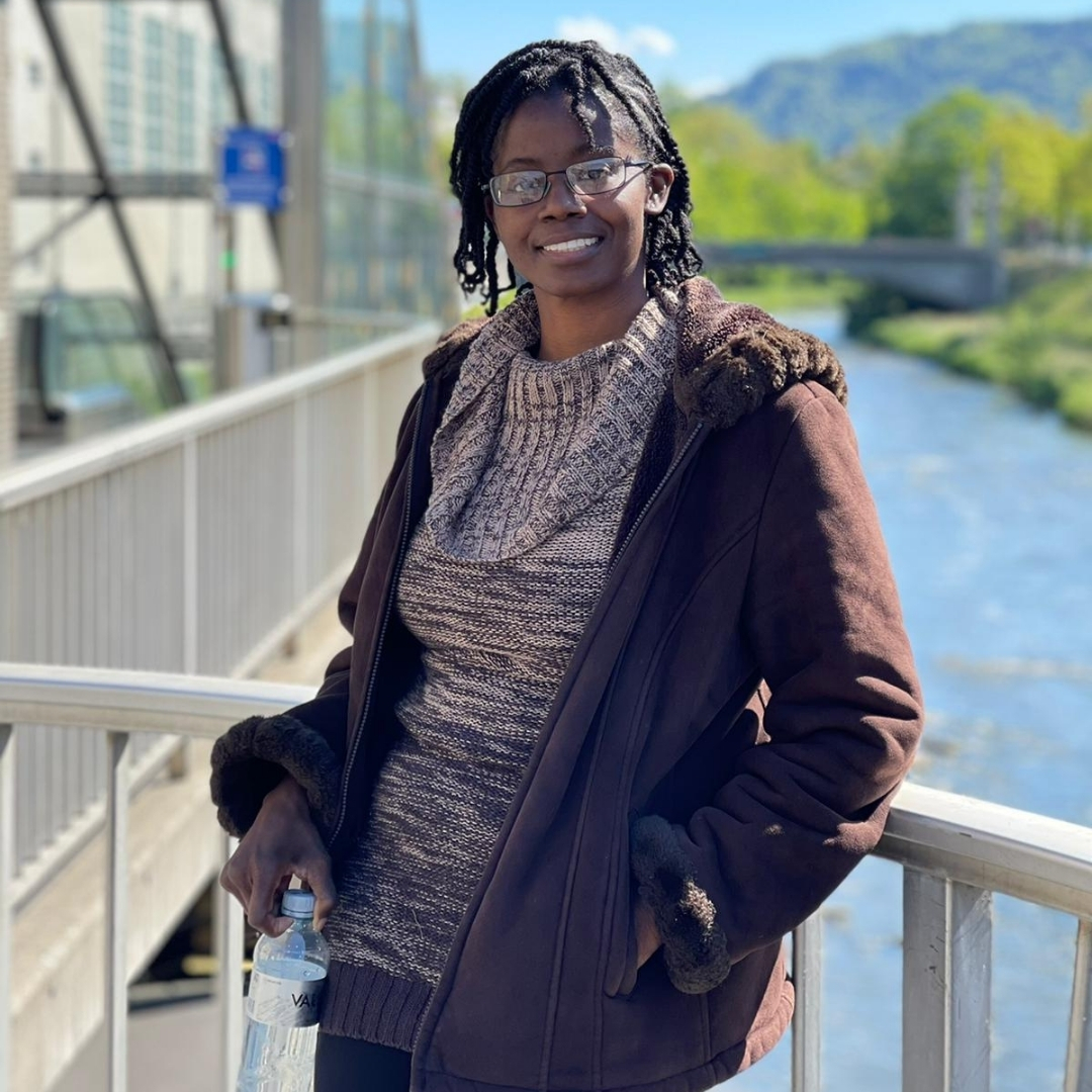 Alt text: Natasha Lewis stands on a bridge overlooking a calm river, wearing a brown coat with a fur-lined hood and a textured knit sweater. Behind her, green trees and rolling hills stretch beneath a blue sky with scattered clouds. The bridge’s metal railings and a nearby modern building with large glass windows blend urban and natural elements in the peaceful outdoor setting.