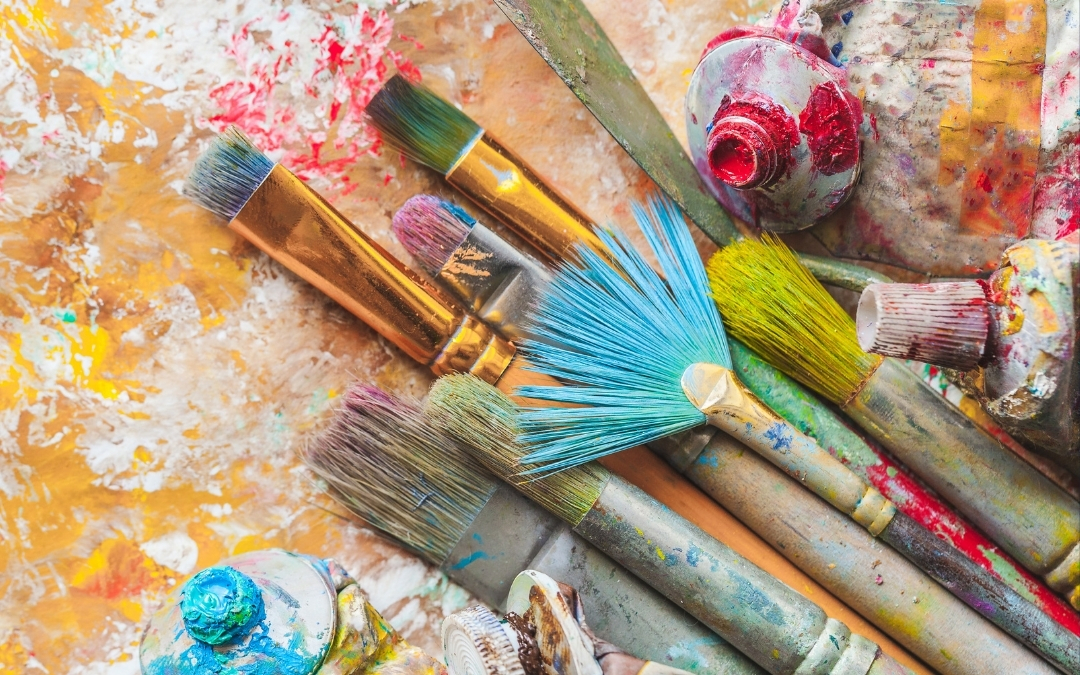 Alt text: A collection of well-used paintbrushes and crumpled paint tubes scattered across a surface splattered with vibrant colors. The brushes vary in size and shape—flat, round, and fan-shaped—while the background features bold strokes of yellow, red, and white, evoking the energy and texture of an artist’s studio.