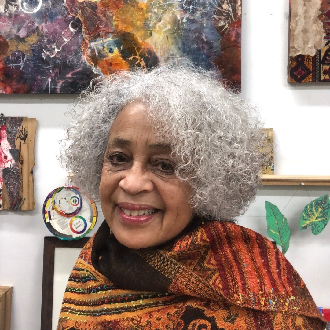Sharon Robinson Portrait of Sharon Robinson smiling in a creative studio space. She has curly gray hair and wears a multicolored shawl in warm tones of red, orange, and brown. Behind her are various artworks, including abstract paintings, a circular mixed‑media piece, and leaf‑shaped decorations.