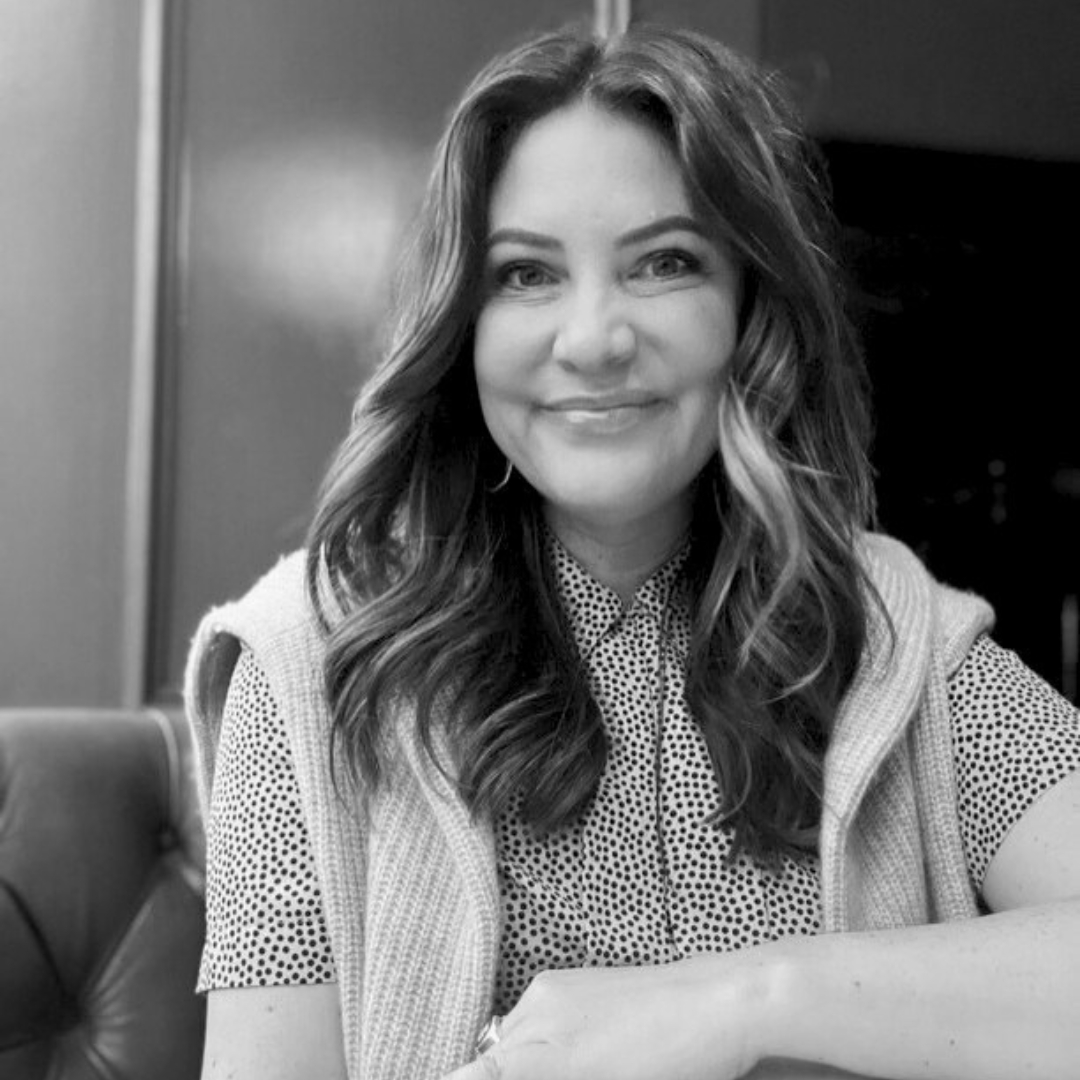 Black‑and‑white portrait of Becca Keaty smiling while seated indoors. She has long wavy hair and wears a patterned button‑up shirt with a sleeveless knit layer over her shoulders. A cushioned chair and plain background sit softly out of focus behind her.