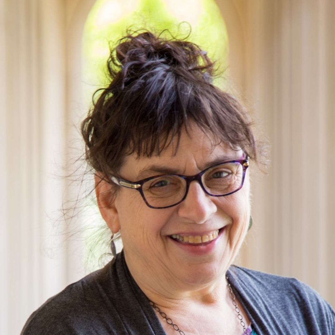 Caryn Mirriam‑Goldberg — Author Portrait Color portrait of author Caryn Mirriam‑Goldberg smiling, wearing glasses and earrings, with short dark hair. Soft natural light filters through vertical blinds in the background.