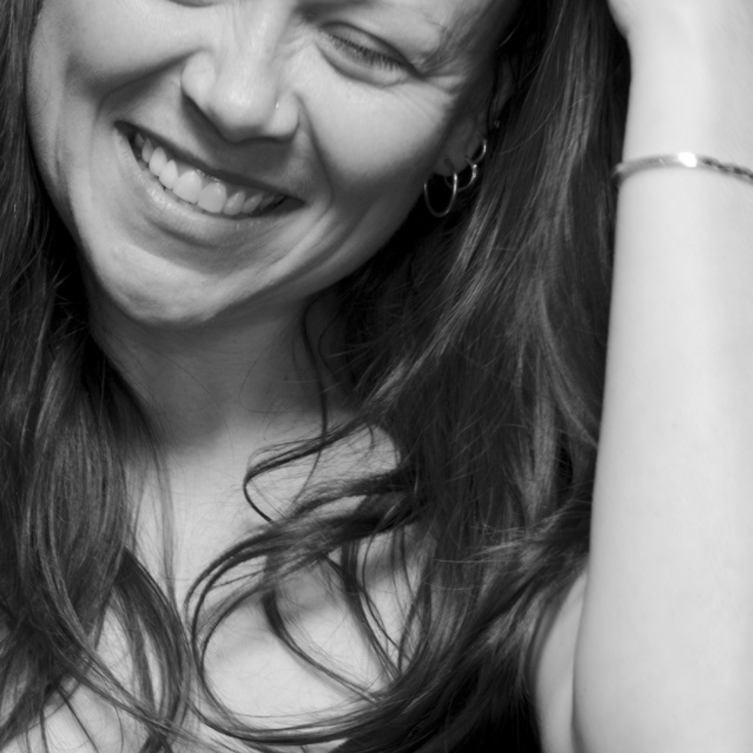 Jeanann Verlee — Author Portrait Black‑and‑white close‑up portrait of poet Jeanann Verlee smiling with her eyes closed. Her long hair falls to one side as she lifts a hand toward it, showing hoop earrings and a bracelet.
