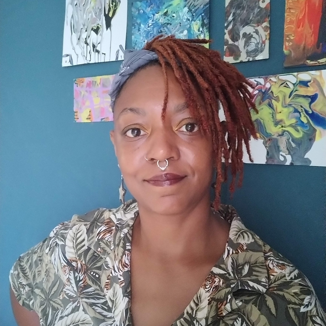 Raychelle Heath — Artist Portrait Color portrait of artist and facilitator Raychelle Heath standing in front of a blue wall with abstract artwork. She has reddish‑brown locs, a patterned headband, a nose ring, and wears a green and brown foliage‑patterned shirt while looking directly at the camera.