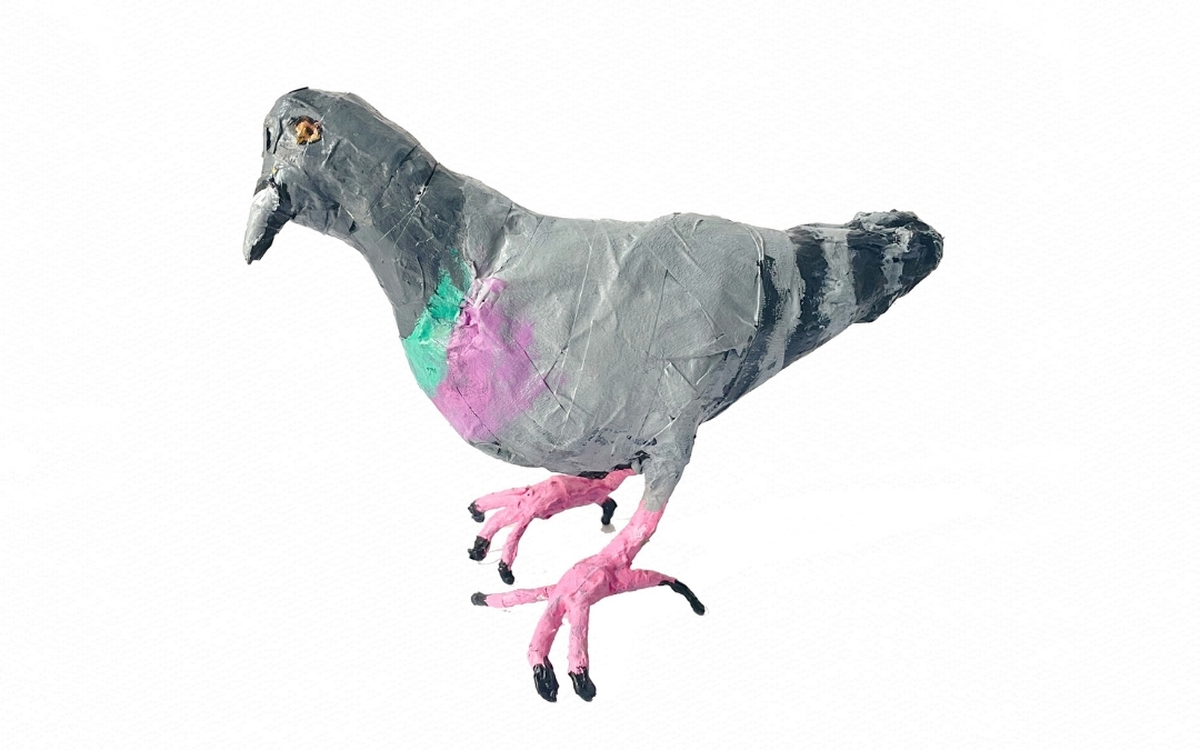 Color photo of a handmade pigeon sculpture created by artist Zehra Khan. The bird is crafted with textured materials and painted in shades of gray with a green and purple patch on its chest, standing on pink legs with black claws. The style is whimsical and expressive, highlighting the playful character of the artwork.