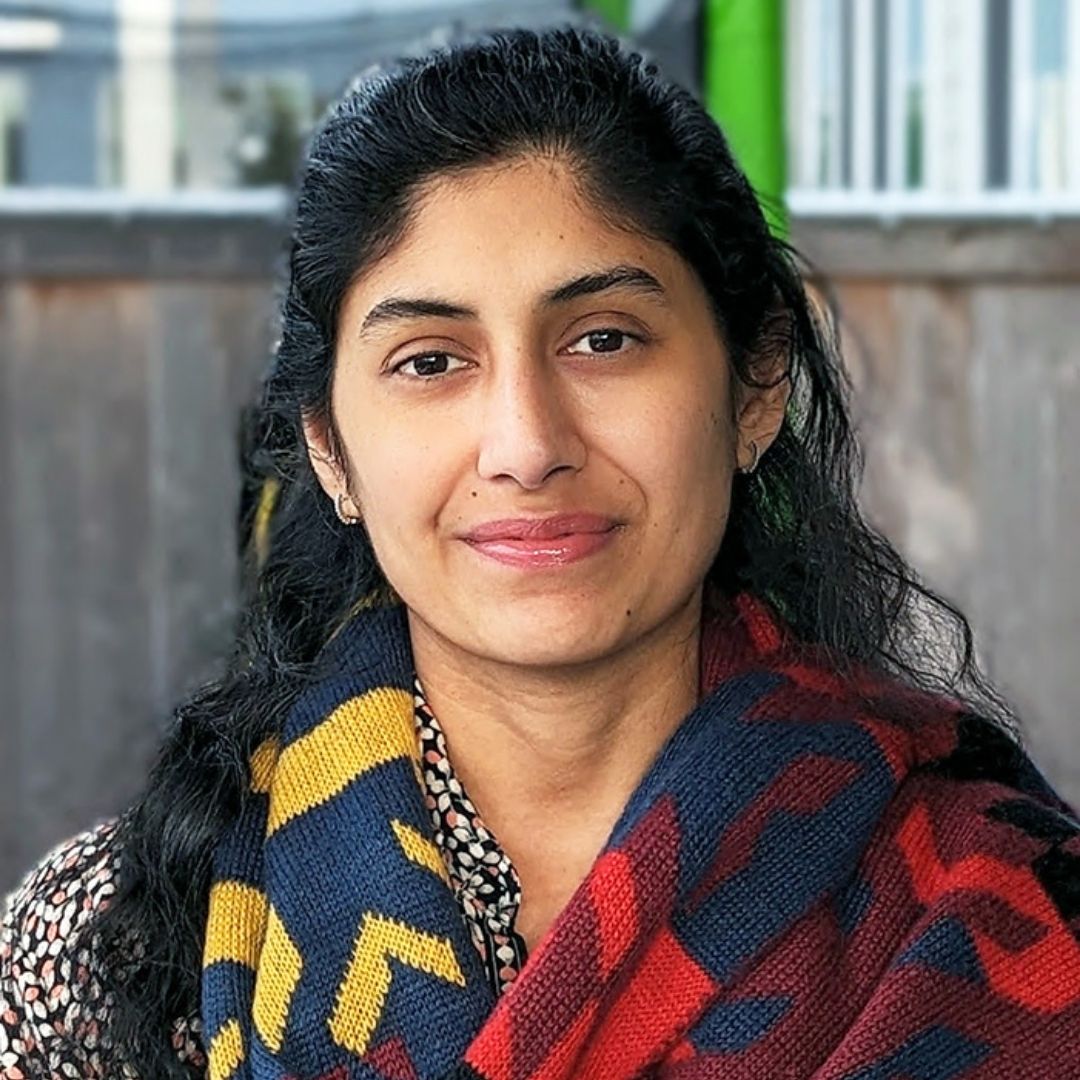 Garima Naredi — Artist Portrait Outdoor portrait of artist Garima Naredi wearing glasses, a patterned shirt, and a colorful knitted scarf with red, blue, and yellow geometric designs. She has long dark hair and is standing in front of a wooden fence and building structure.