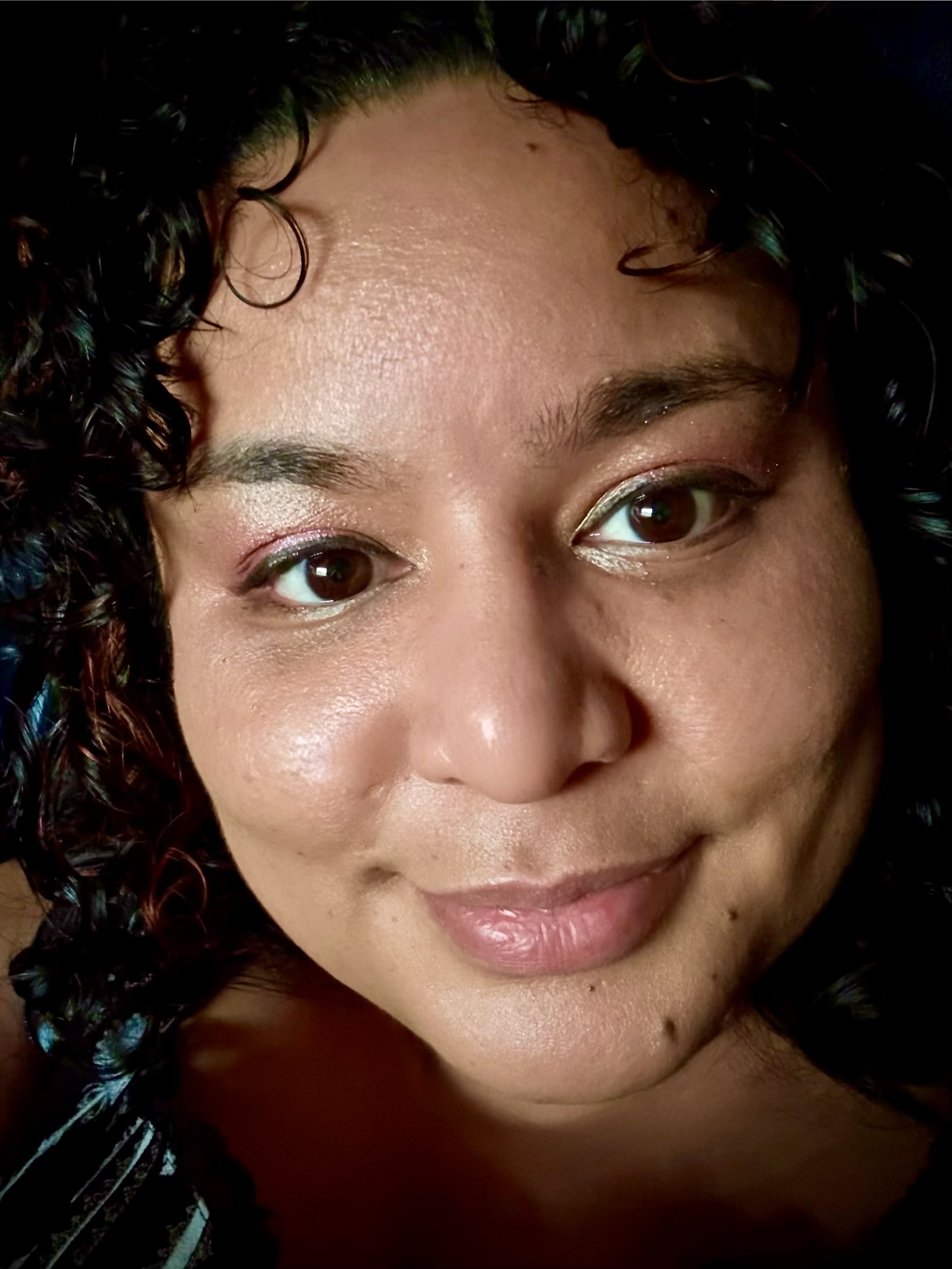 Close‑up portrait of artist Belena Stuart Marquez with curly dark hair and brown eyes, softly lit against a dark background. Her face is centered and in sharp focus.