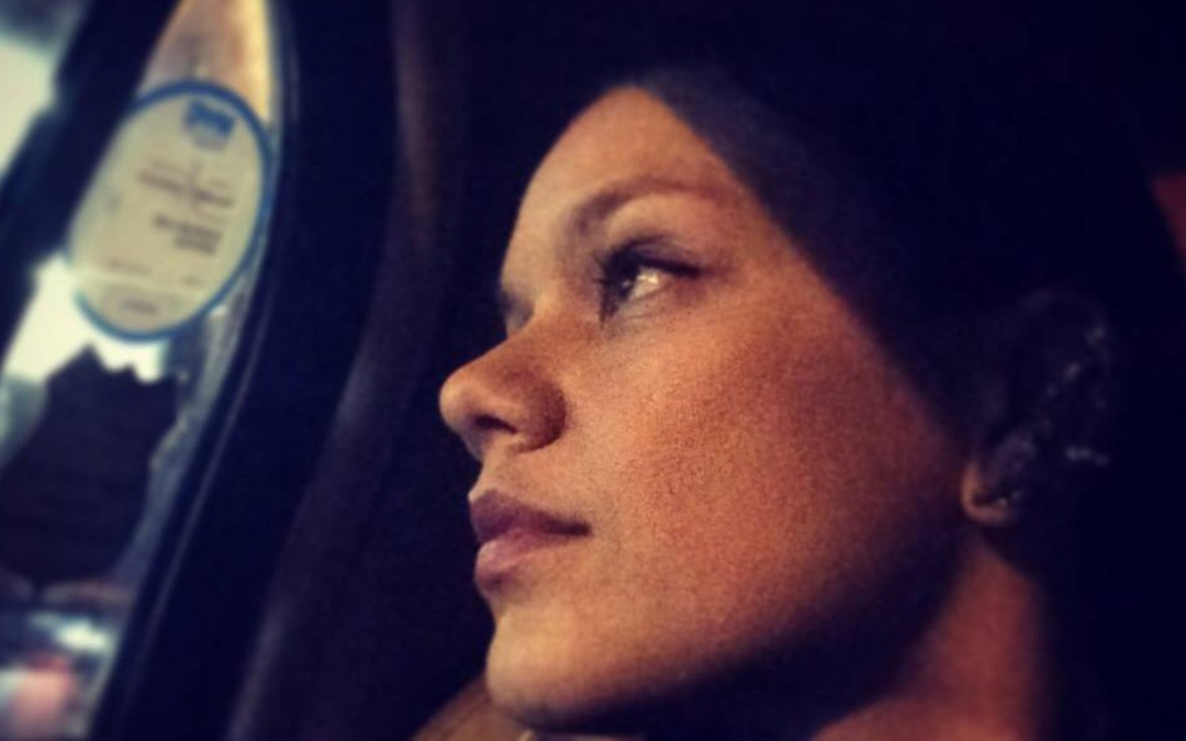 A side‑profile portrait of Christina Olivares looking out a window at night, softly illuminated by ambient light. The image captures a contemplative expression and warm tones against a dark background.