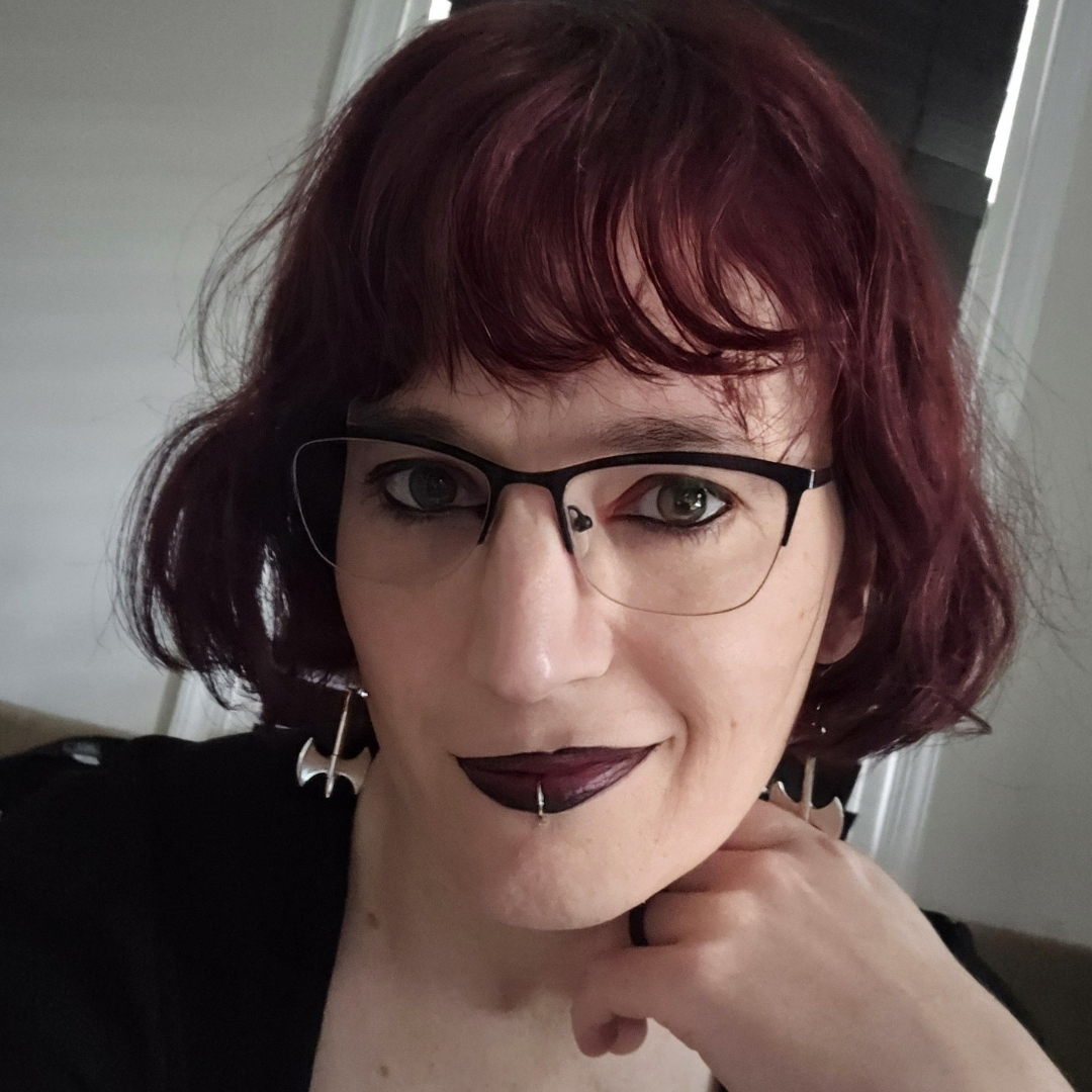Cathrine Schmid — Author Portrait Color portrait of Cathrine Schmid indoors, resting her chin on her hand. She has short wavy burgundy hair, glasses, dark lipstick, a lip piercing, and bat‑shaped earrings. A window with closed blinds and a white wall appear softly in the background.