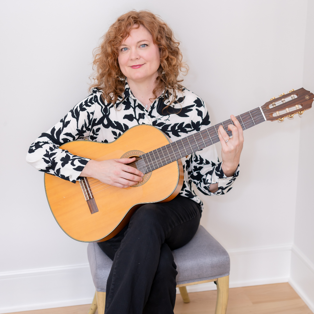 Maureen Andary — Portrait — 1080×1080 — 2026 Musician Maureen Andary plays or poses with an acoustic guitar while wearing a white shirt with a black leaf pattern, seated against a soft, light background.