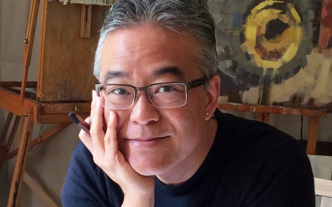 Aaron Caycedo‑Kimura rests his face on one hand while holding a pen, smiling gently. An easel and an abstract circular painting sit behind him, creating a warm, creative studio atmosphere.
