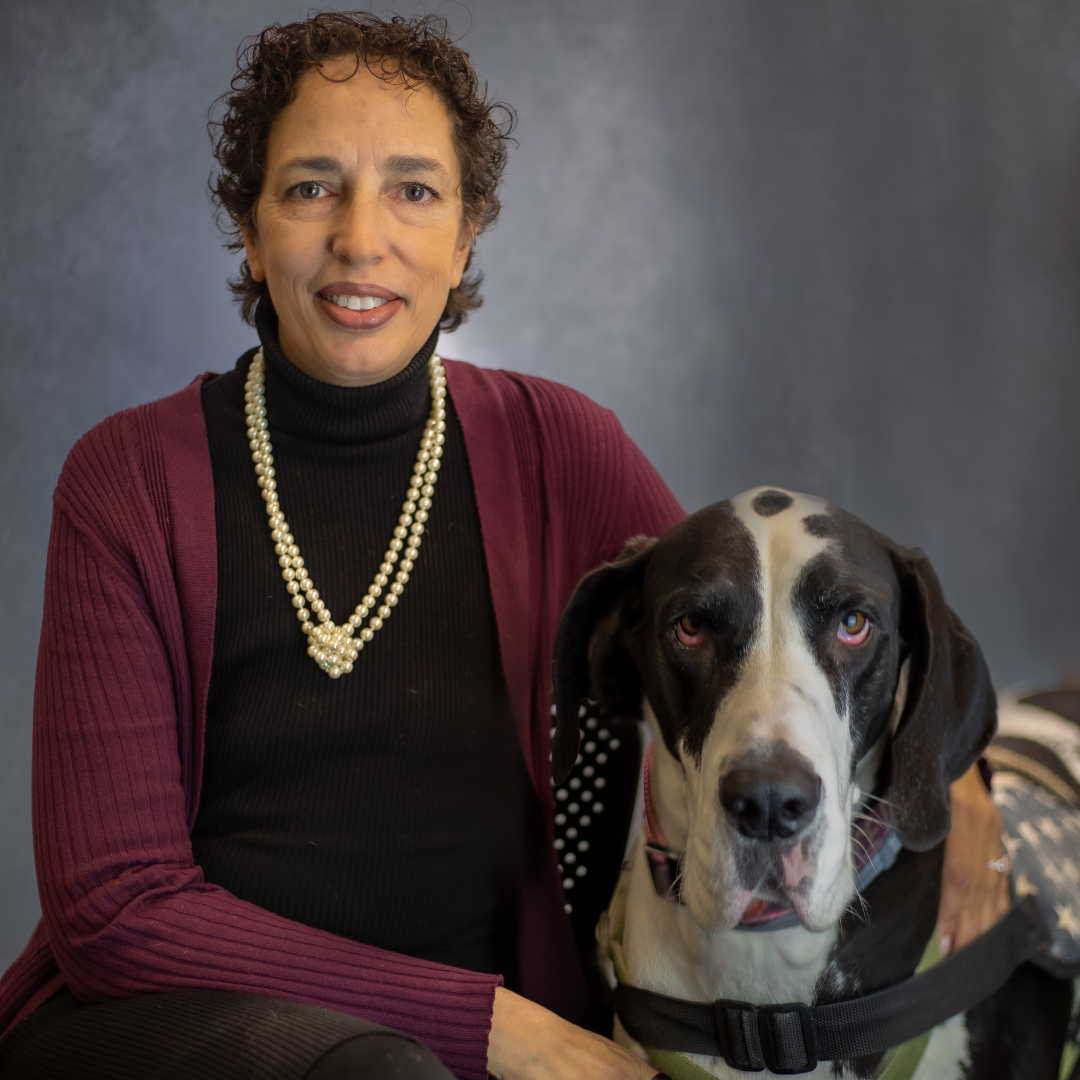 Artist Micaila Britto sits beside her large black‑and‑white Great Dane against a dark gray backdrop. She wears a maroon cardigan, black turtleneck, and double‑strand pearl necklace, smiling warmly while the dog looks forward.