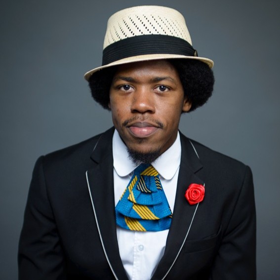 Christylez Bacon wears a straw hat, black suit jacket with white piping, white dress shirt, patterned blue‑and‑yellow necktie, and a red rose lapel pin while posing against a gray background.