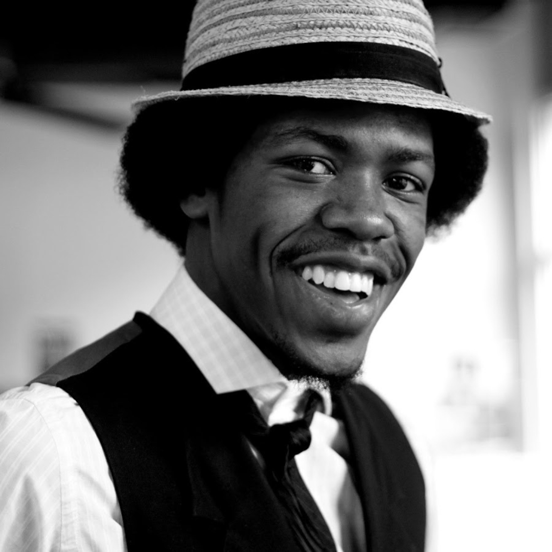 Christylez Bacon — Hip Hop Artist/Instrumentalist Christylez Bacon wears a straw hat, vest, tie, and collared shirt while posing indoors in a softly lit space. The black‑and‑white portrait highlights his expressive features and classic style.