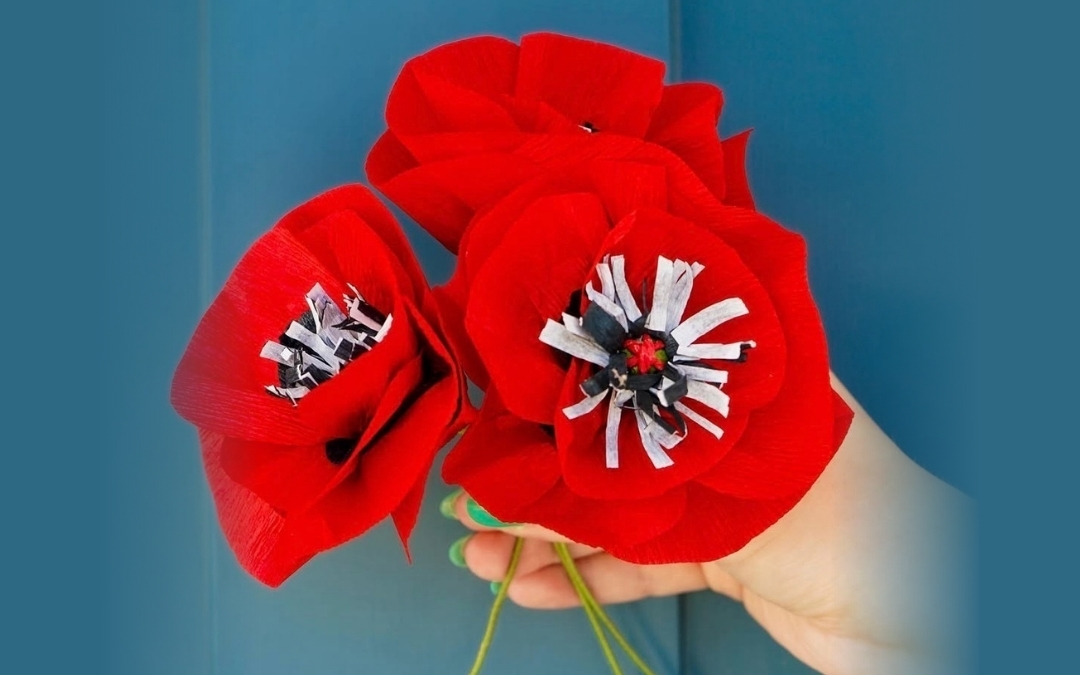 Crepe Paper Flowers — Visual Arts Workshop A hand holds three handmade red crepe paper flowers resembling poppies, each with textured petals and black‑and‑white paper centers. The vivid red blooms stand out against a smooth blue background, highlighting their delicate craftsmanship.