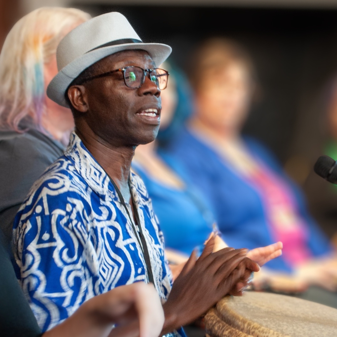 Kofi Dennis - Drummer/Storyteller Kofi Dennis sits playing a hand drum while wearing a light gray hat, glasses, and a blue patterned shirt. People seated in the background suggest a group drumming or cultural activity.