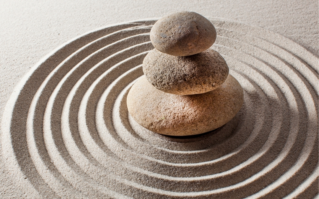 Meditation — Mindfulness Workshop Three smooth stones are stacked in a balanced tower on fine sand, surrounded by concentric circular patterns raked into the surface, creating a calm, Zen‑inspired scene.