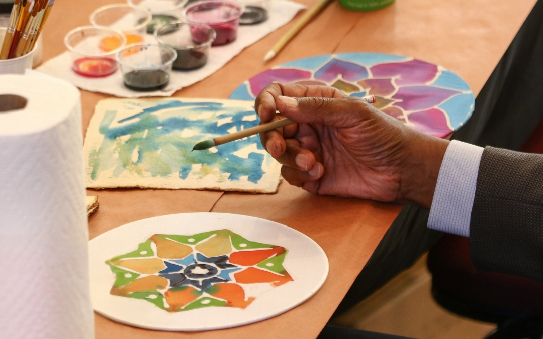 Painting — Visual Arts Workshop A person paints blue and green watercolor strokes on paper at a table surrounded by small paint cups, paper towels, and two colorful circular mandala‑style artworks.