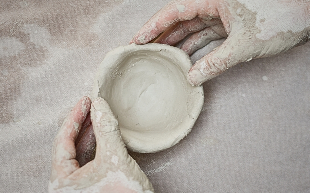 Paper Clay — Visual Arts Workshop Two clay‑covered hands hold a small, round, hand‑shaped clay bowl over a light surface dusted with clay powder. The image captures the tactile, creative process of forming pottery by hand.
