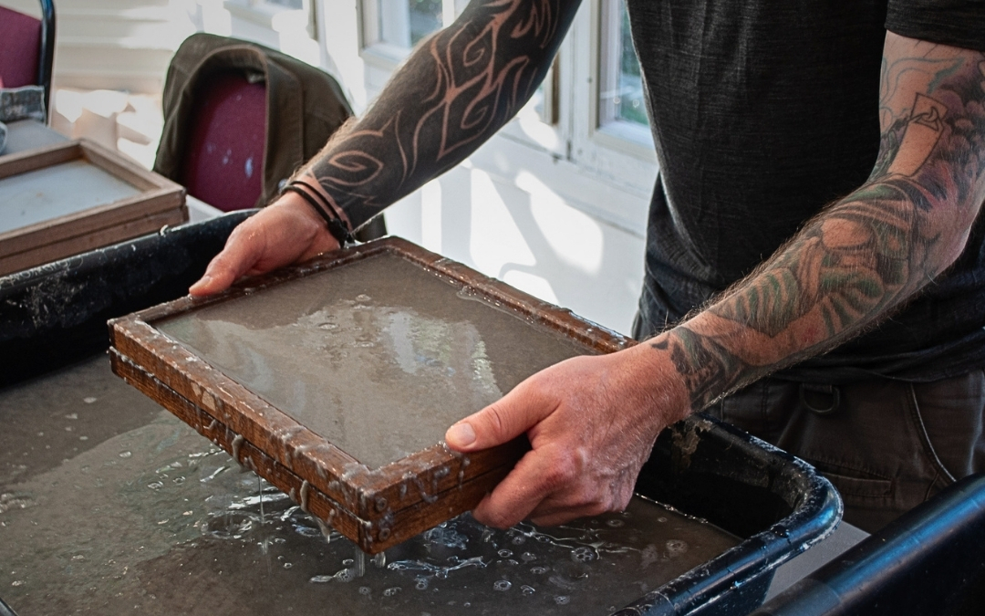 Papermaking — Visual Arts Workshop Tattooed hands lift a wooden deckle and mold from a basin of water, revealing a thin layer of pulp that will become handmade paper. The image captures the tactile, creative process of traditional papermaking.