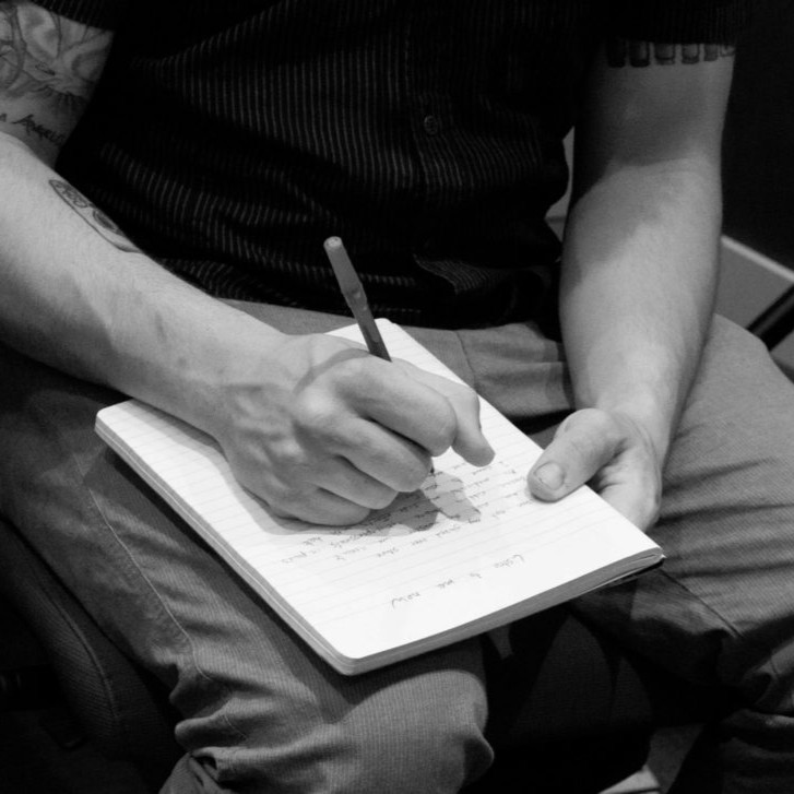 Creative Writing Workshop — Writing Photo (B&W) A black‑and‑white photo shows a tattooed person seated and writing in a lined notebook with a pencil. The notebook rests on their lap, capturing a moment of focused, expressive writing that conveys creativity and reflection.