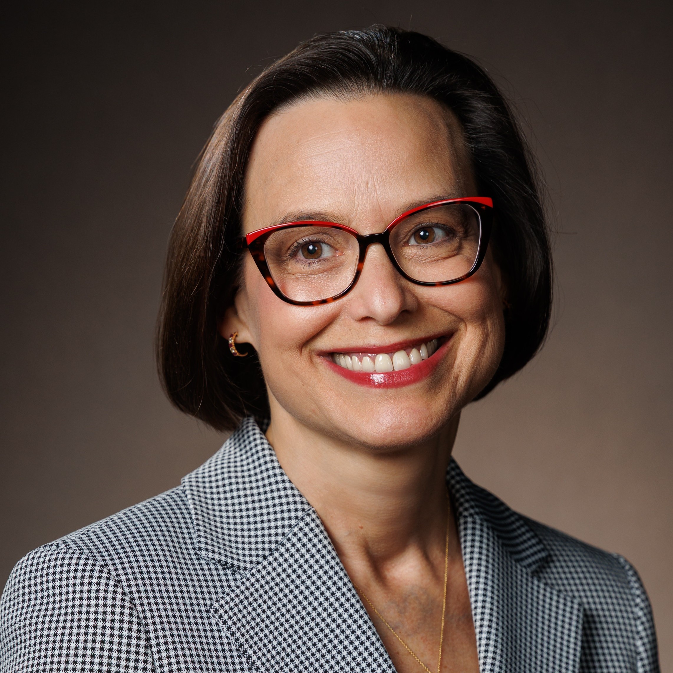 COL (ret) Jessica Bunin, MD, MHPE — Headshot COL (ret) Jess Bunin, MD, MHPE smiles warmly while wearing red‑and‑brown glasses, a gray checkered blazer, and a gold necklace. She has short dark hair and is photographed against a soft brown gradient background.