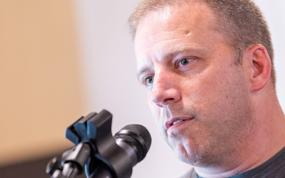 A close‑up portrait of Ben Weakley speaking into a professional microphone, with short hair, a focused expression, and a softly blurred indoor background.